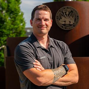 Headshot of a man with brown hair wearing a grey shirt standing outside on a sunny day with his arms crossed 